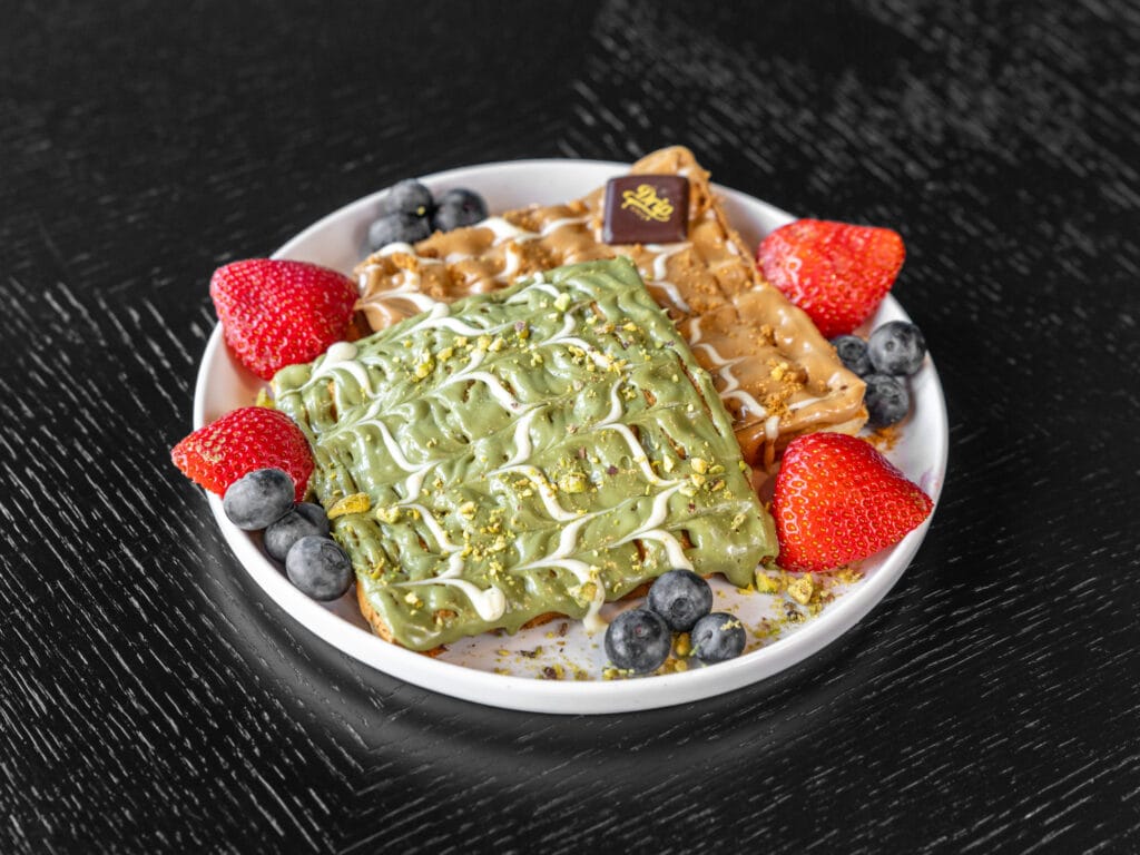 Drip Cafe Belgian Waffles, served in two pieces, one covered in pistachio drizzle and the other in caramel drizzle, garnished with fresh strawberries and blueberries.