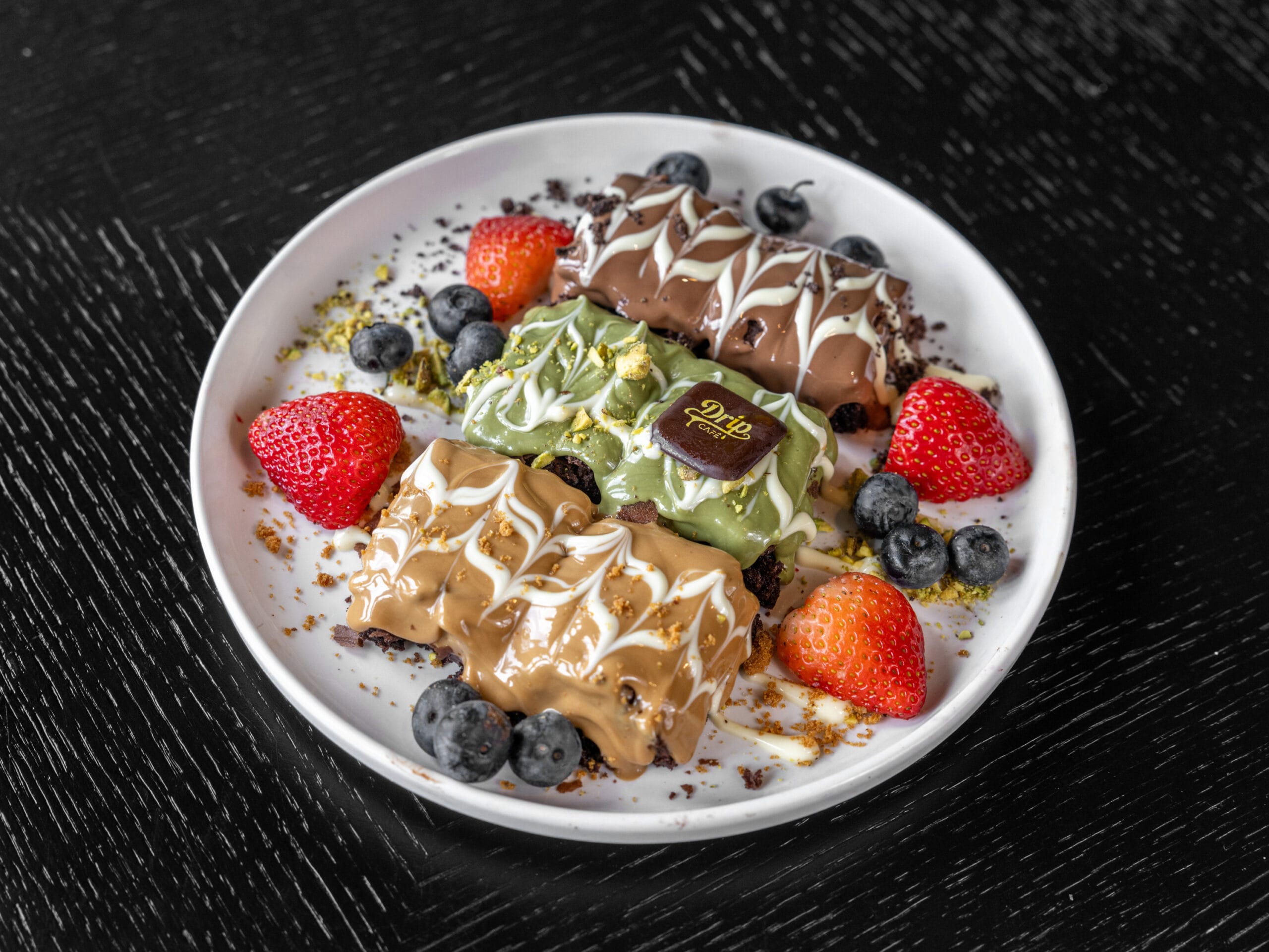 Drip Cafe Brownie dessert plate featuring three rich rectangular brownie pieces, topped with white chocolate, pistachio, and caramel drizzles, served with fresh strawberries and blueberries.