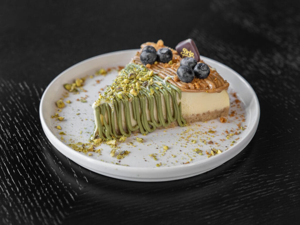 Drip Cafe Cheesecake slice with a two-tone topping of pistachio cream drizzle and caramel/cookie butter, garnished with fresh blueberries, pistachio crumbs, and a Drip Cafe chocolate piece.