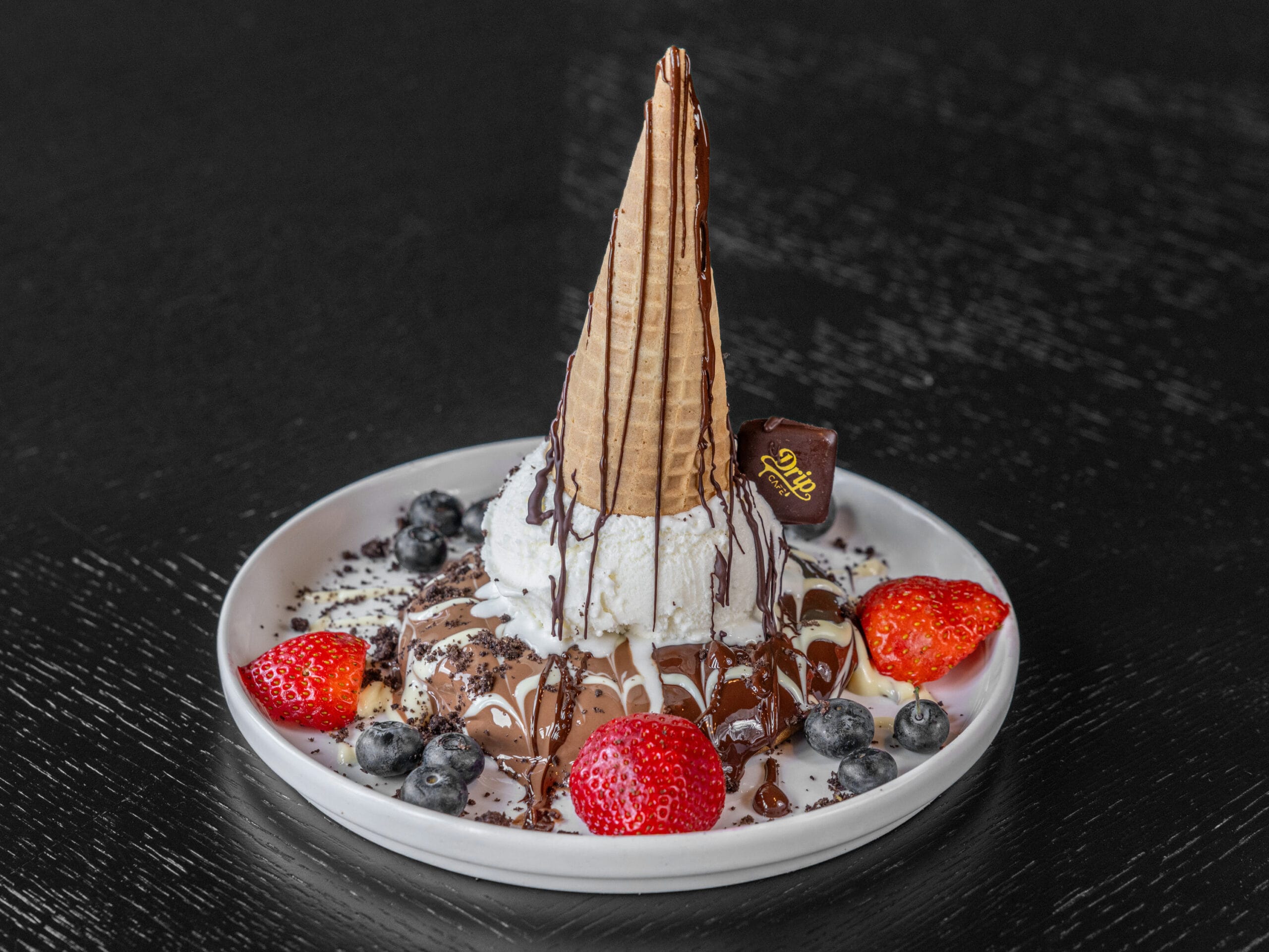 Drip Cafe Drip Drop Cookie dessert, featuring a vanilla ice cream scoop topped with a chocolate-drizzled waffle cone, resting on a bed of melted chocolate, cookie crumbs, fresh strawberries, and blueberries.