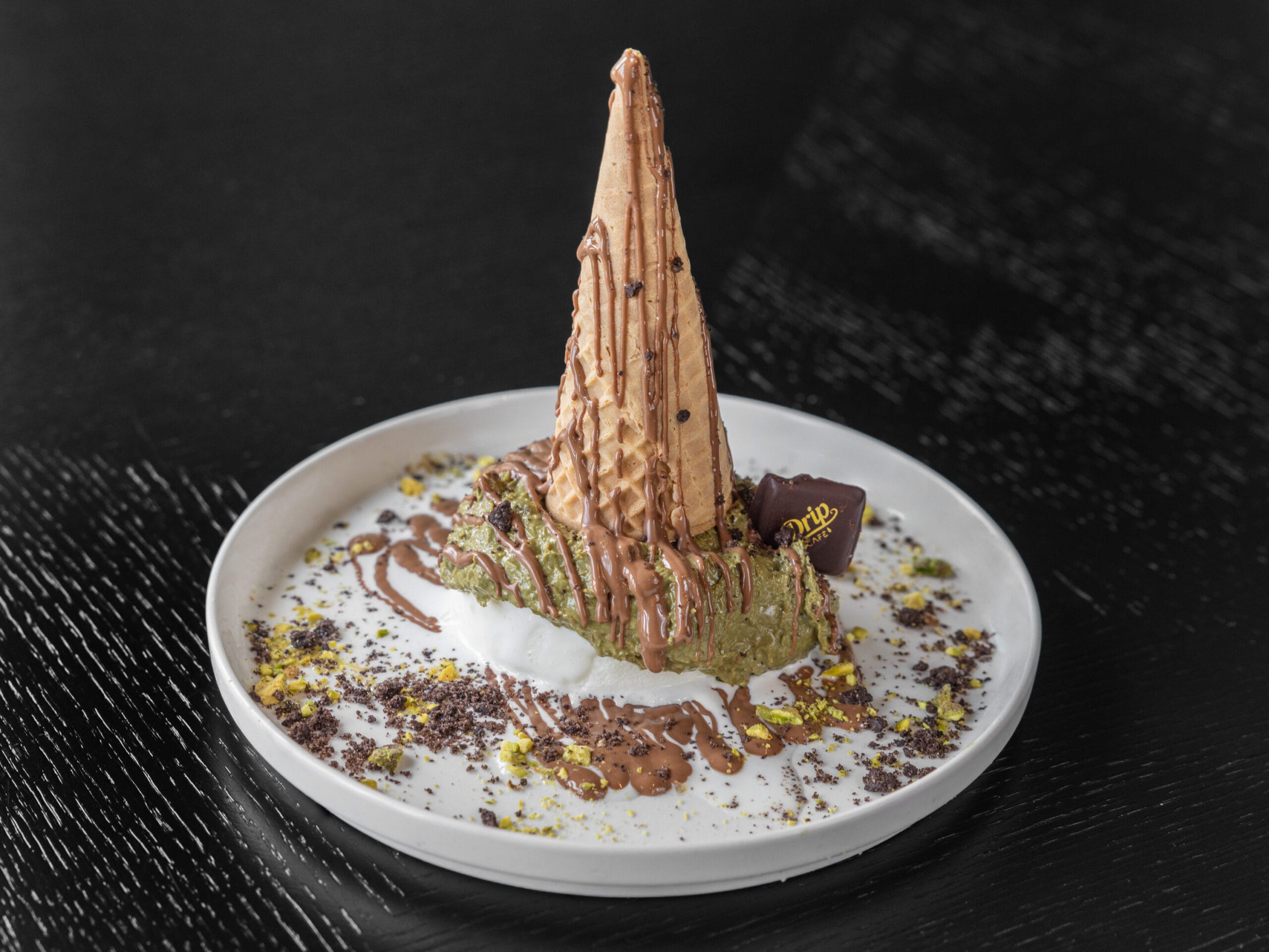 Drip Cafe Kunafa Cone, a tower dessert with crispy kunafa and pistachio cream on a bed of sauce and cookie crumbs, crowned with a chocolate-drizzled cone.