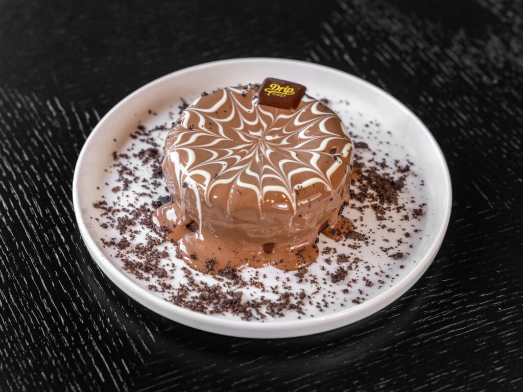 Drip Cafe Lava Cake, a single-serving chocolate cake coated in melted chocolate, topped with a white drizzle design, cookie crumbs, and a Drip Cafe chocolate piece.
