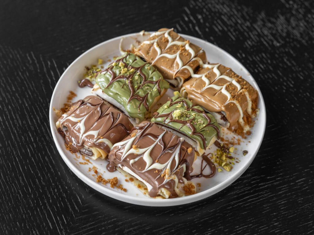 Drip Cafe Sushi Crepes dessert plate featuring multiple pieces of rolled crepes topped with milk chocolate, pistachio, and cookie butter sauces, and garnished with crumbs and pistachio dust.