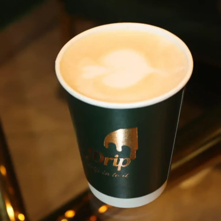 A creamy cappuccino with heart-shaped latte art served in a dark green Drip cafe cup with gold lettering, highlighting one of the signature Drip cafe menu items.