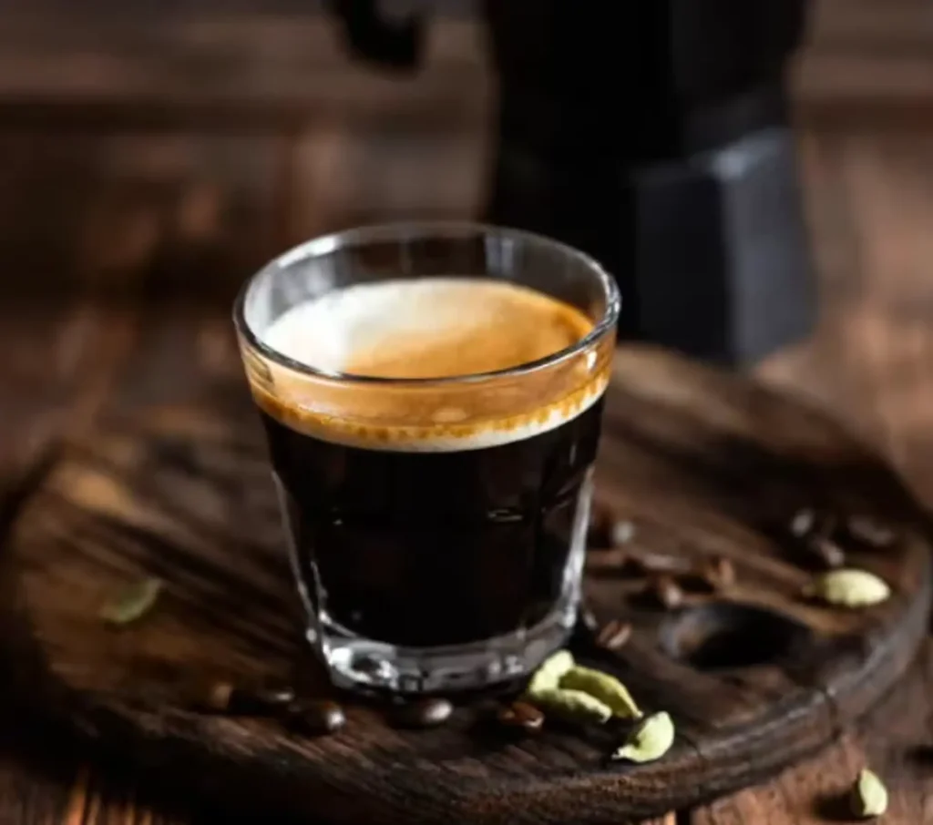 A freshly brewed espresso served in a small glass cup on a rustic wooden board, surrounded by coffee beans and cardamom pods, featured on the Drip cafe menu.