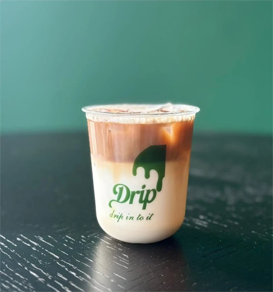 Drip Cafe Spanish Latte, a layered iced coffee made with sweetened milk and espresso in a branded Drip cup, set against a green background.